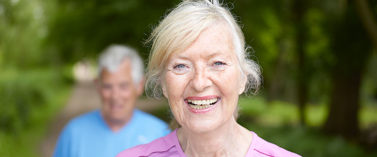 Sprek eldre dame som smiler på tur i front. Eldre mann i bakgrunnen. 