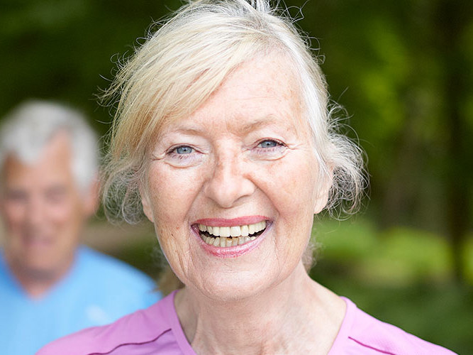 Sprek eldre dame som smiler på tur i front. Eldre mann i bakgrunnen.